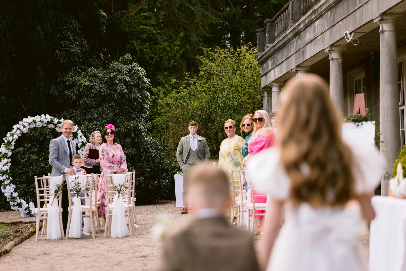 A group of well-dressed people stand and sit outdoors near a decorated arch and building, watching two children walk toward them.