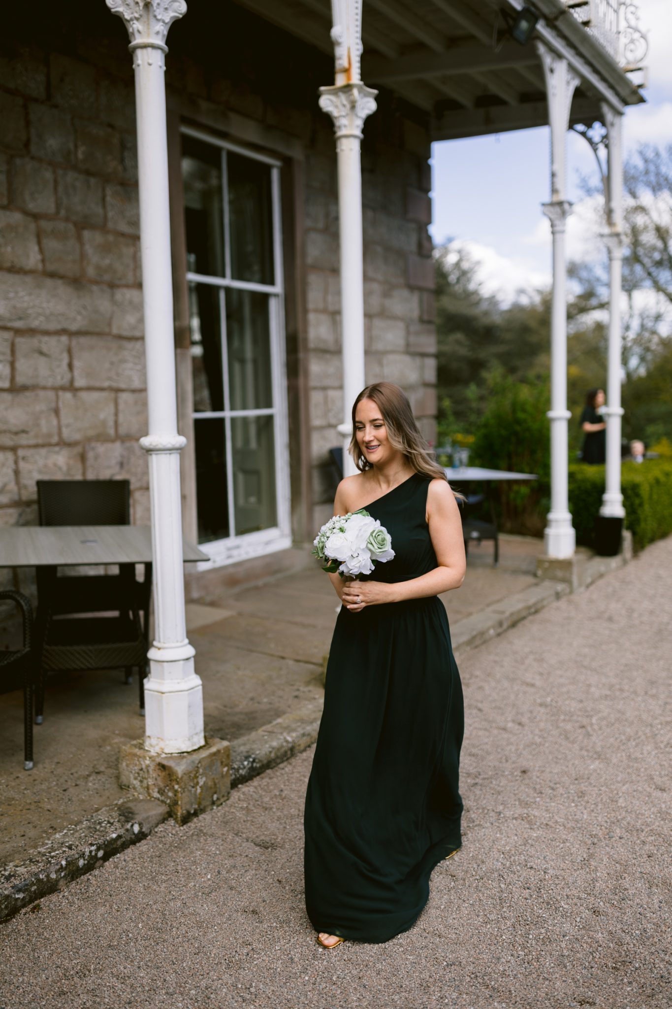 A woman in a long black dress walks outside near a stone building, holding a bouquet of white and green flowers.