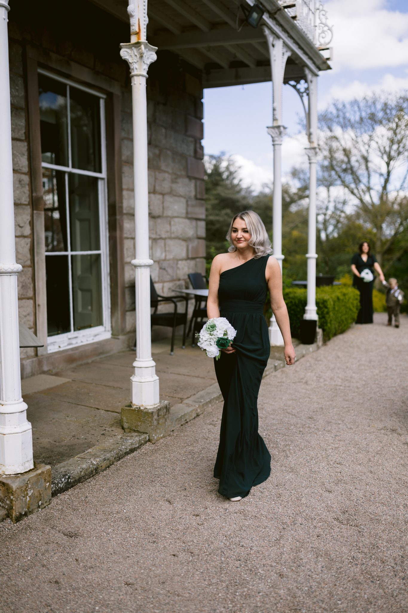 A woman in a dark green one-shoulder dress holds a bouquet of white flowers while walking outside a stone building with white columns.