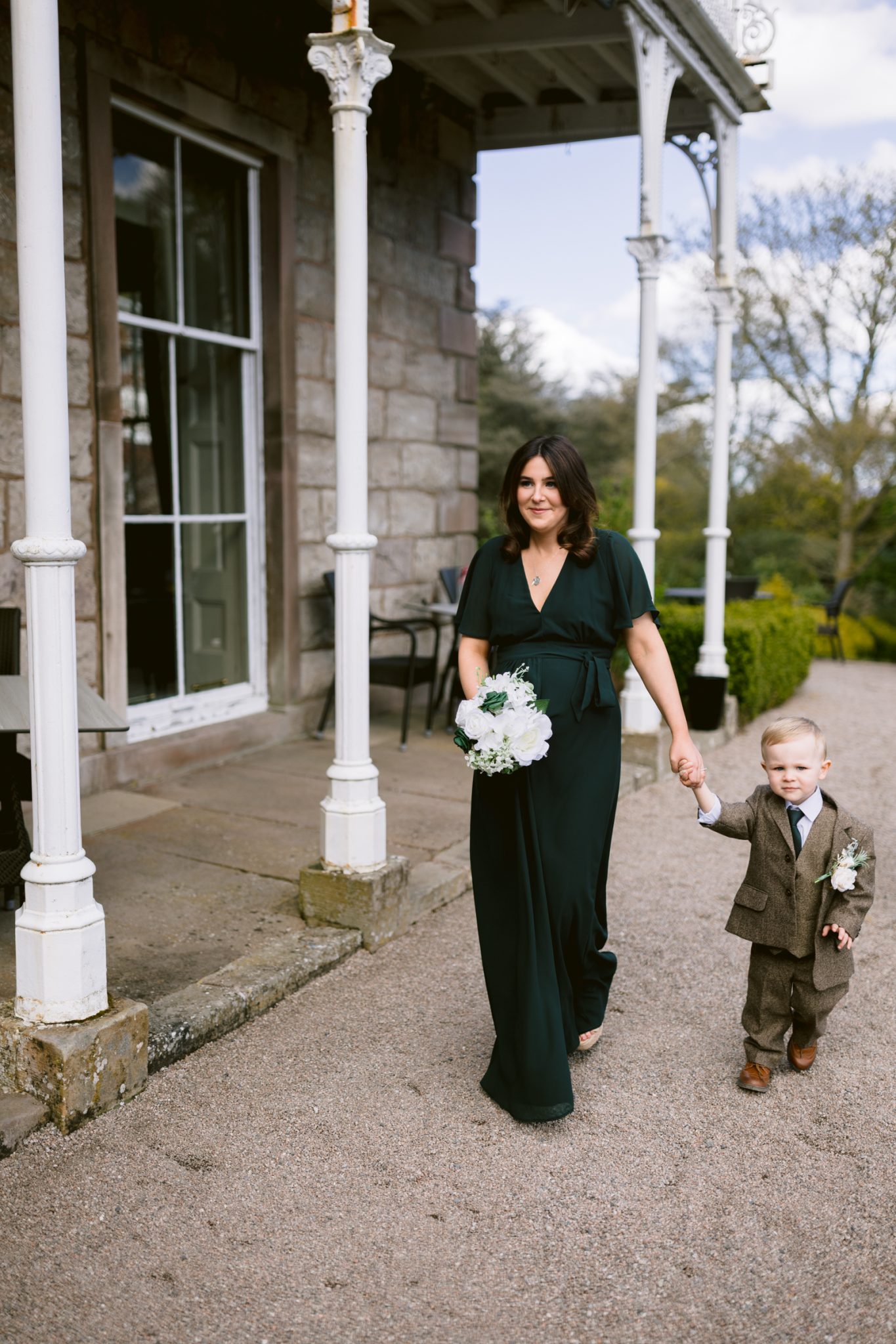 A woman in a dark green dress holds a bouquet and walks hand in hand with a young boy in a suit outside a stone building.