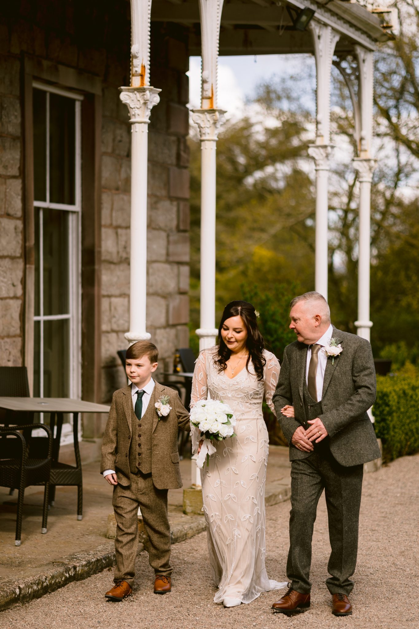 A bride in a white dress walks outdoors, holding a bouquet, accompanied by a man and a young boy in suits on a gravel path beside a building.