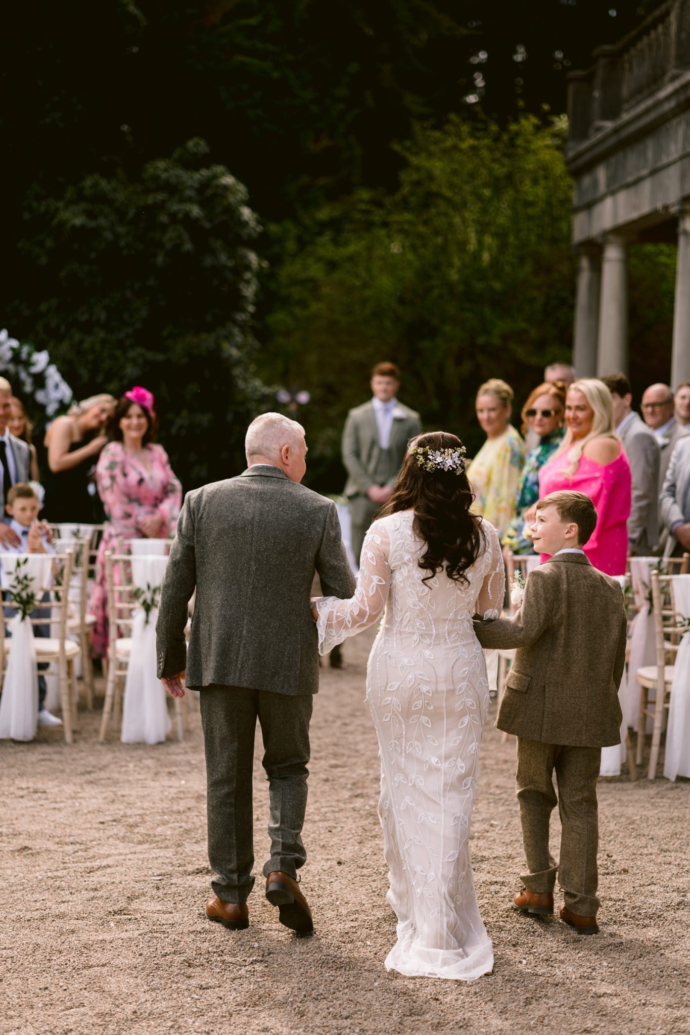 A bride in a white dress walks down the aisle outdoors, accompanied by an older man and a young boy, toward seated guests.
