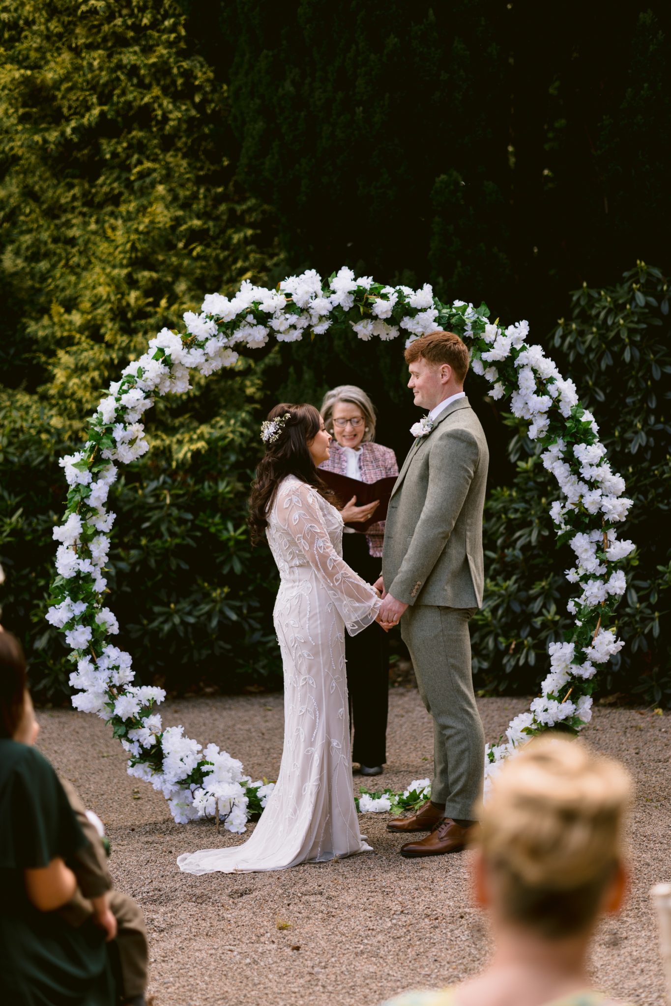 A bride and groom stand holding hands in front of a floral arch during an outdoor wedding ceremony, with an officiant behind them.