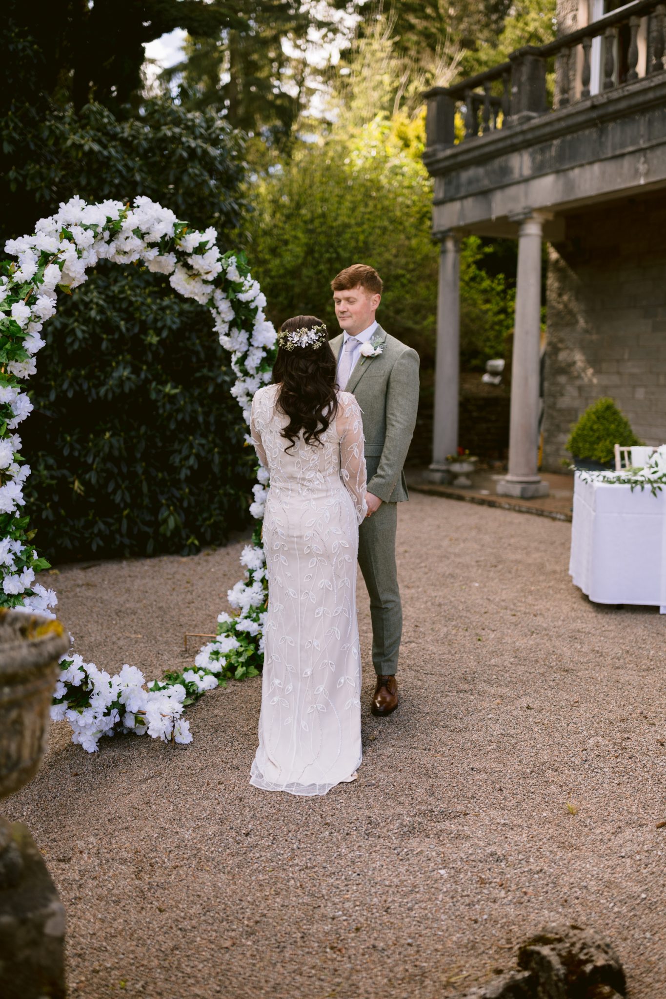 A bride and groom stand facing each other outdoors near a floral arch, with trees and a stone building in the background.