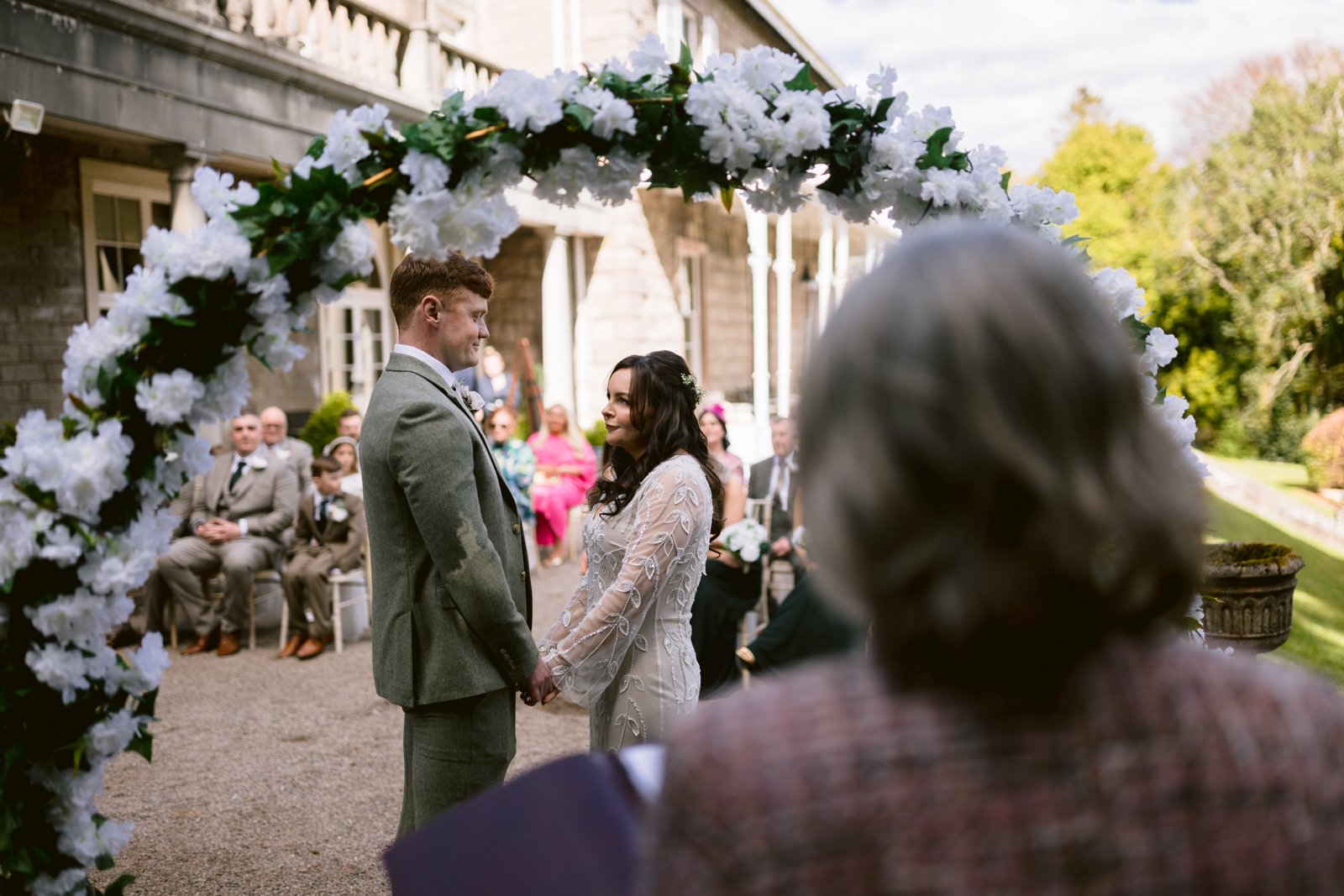 A couple stands under a floral arch, holding hands during an outdoor wedding ceremony, with guests seated and an officiant in the foreground.
