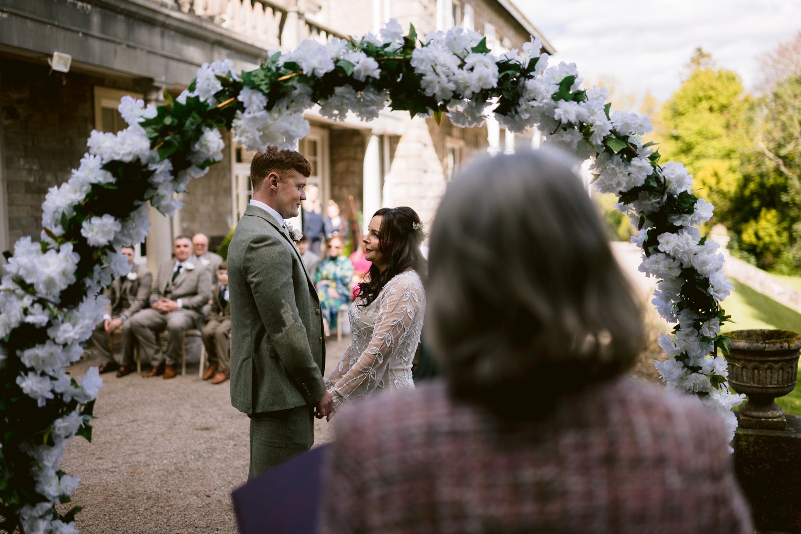 A couple stands under a floral arch holding hands during an outdoor wedding ceremony, with seated guests and an officiant present.