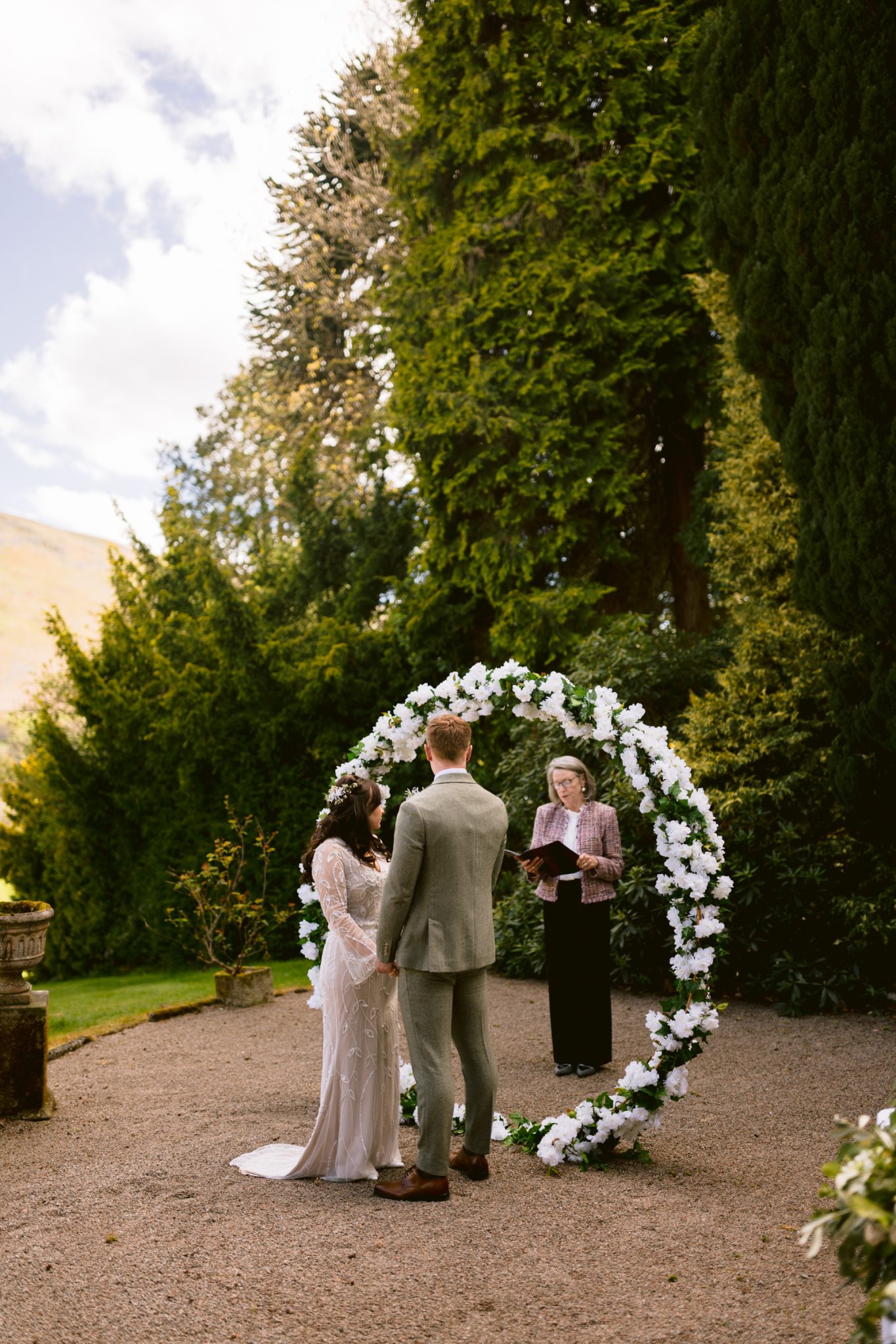 A couple stands facing each other under a circular floral arch outdoors, with an officiant holding a book and leading a wedding ceremony. Lush greenery surrounds the scene.