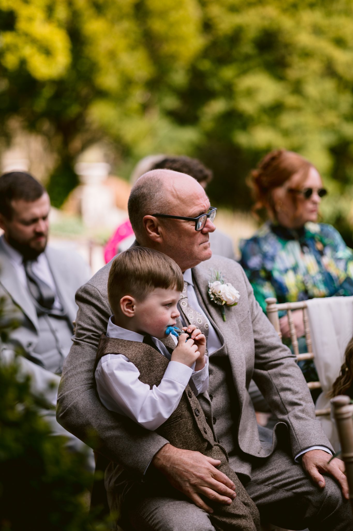 An older man in a suit sits with a young boy on his lap at an outdoor event; both wear formal attire and look ahead, surrounded by other seated guests.