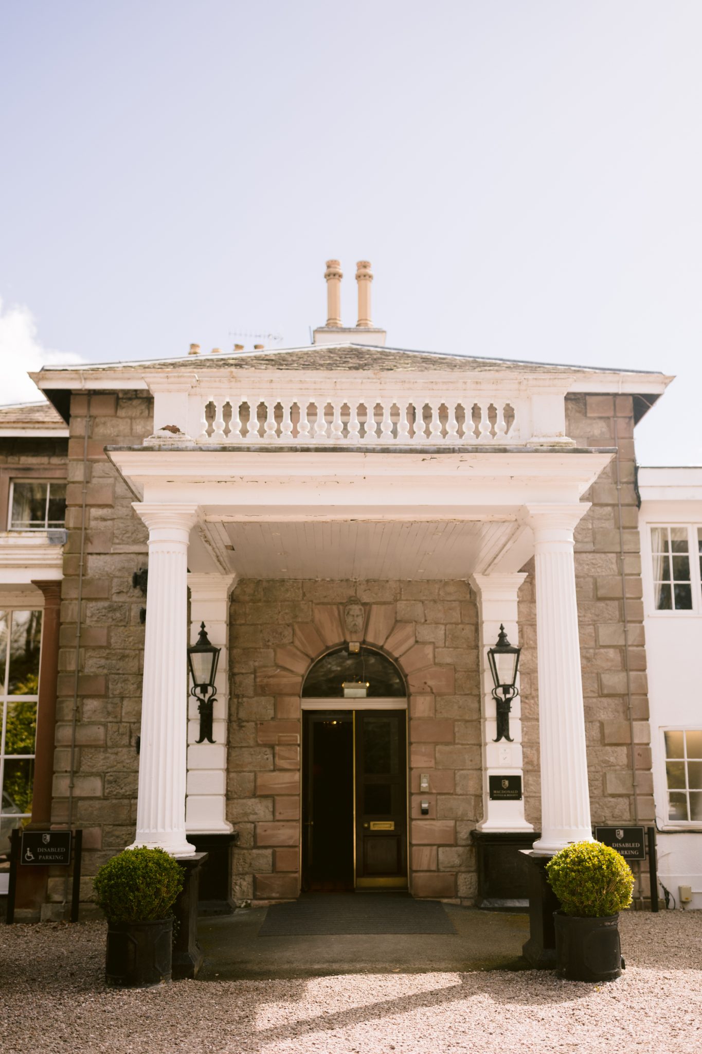 A grand entrance to a stone building with white columns, large lanterns, and potted shrubs on either side of the doorway.