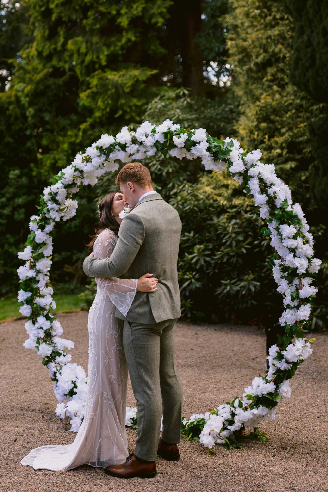 A couple dressed in formal attire kisses in front of a circular floral arch outdoors, surrounded by greenery.