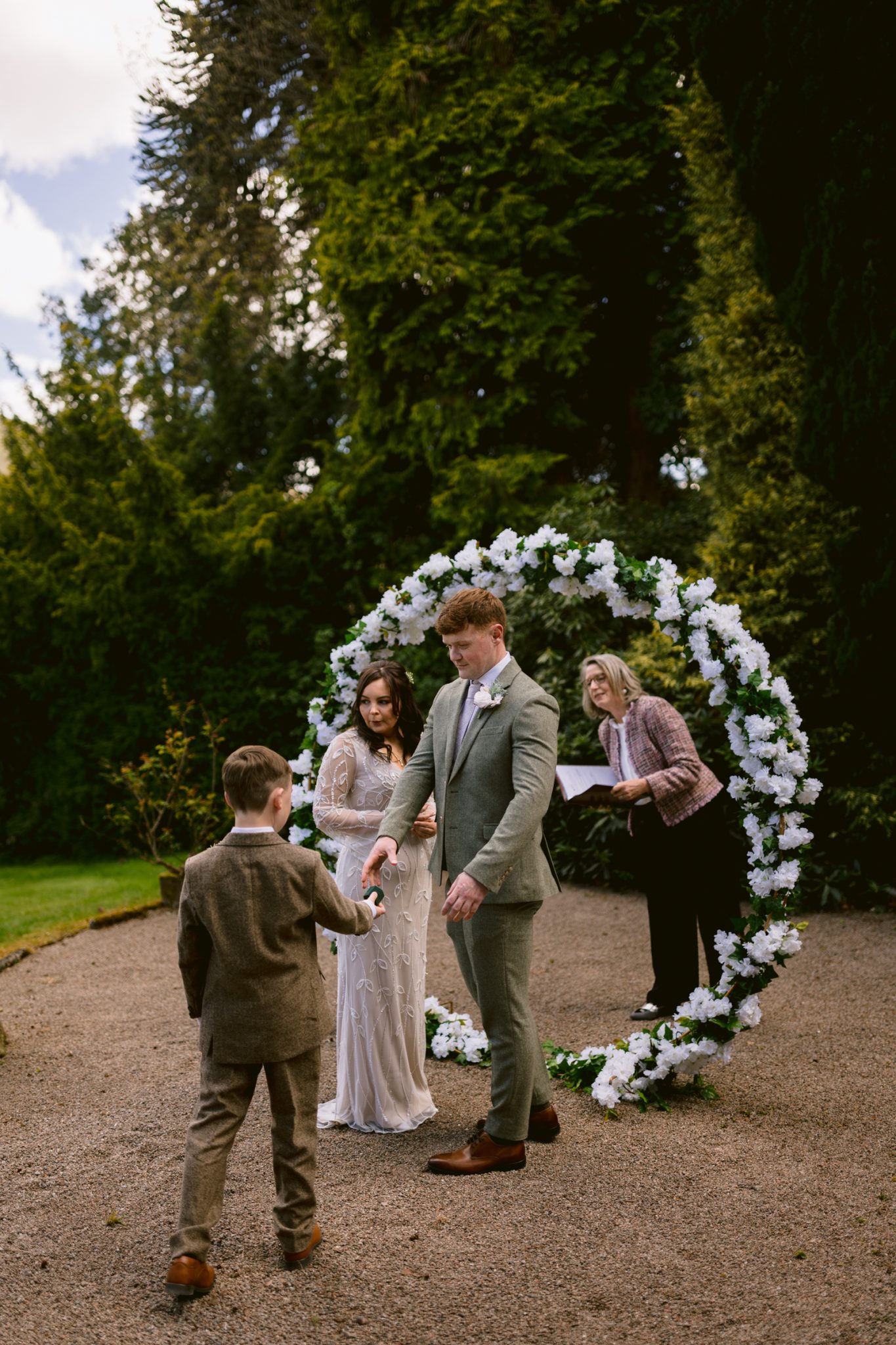 A bride and groom stand in front of a circular floral arch outdoors while a young boy hands something to the groom; an officiant stands behind them holding papers.
