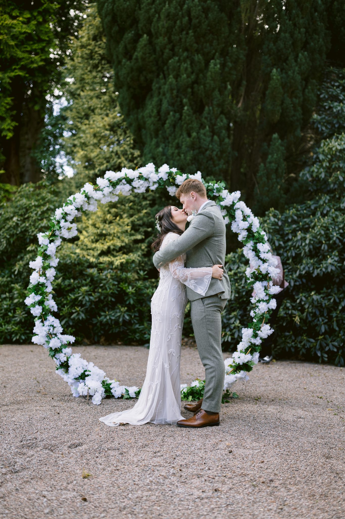 A bride and groom embrace in front of a circular floral arch outdoors, surrounded by greenery and trees.