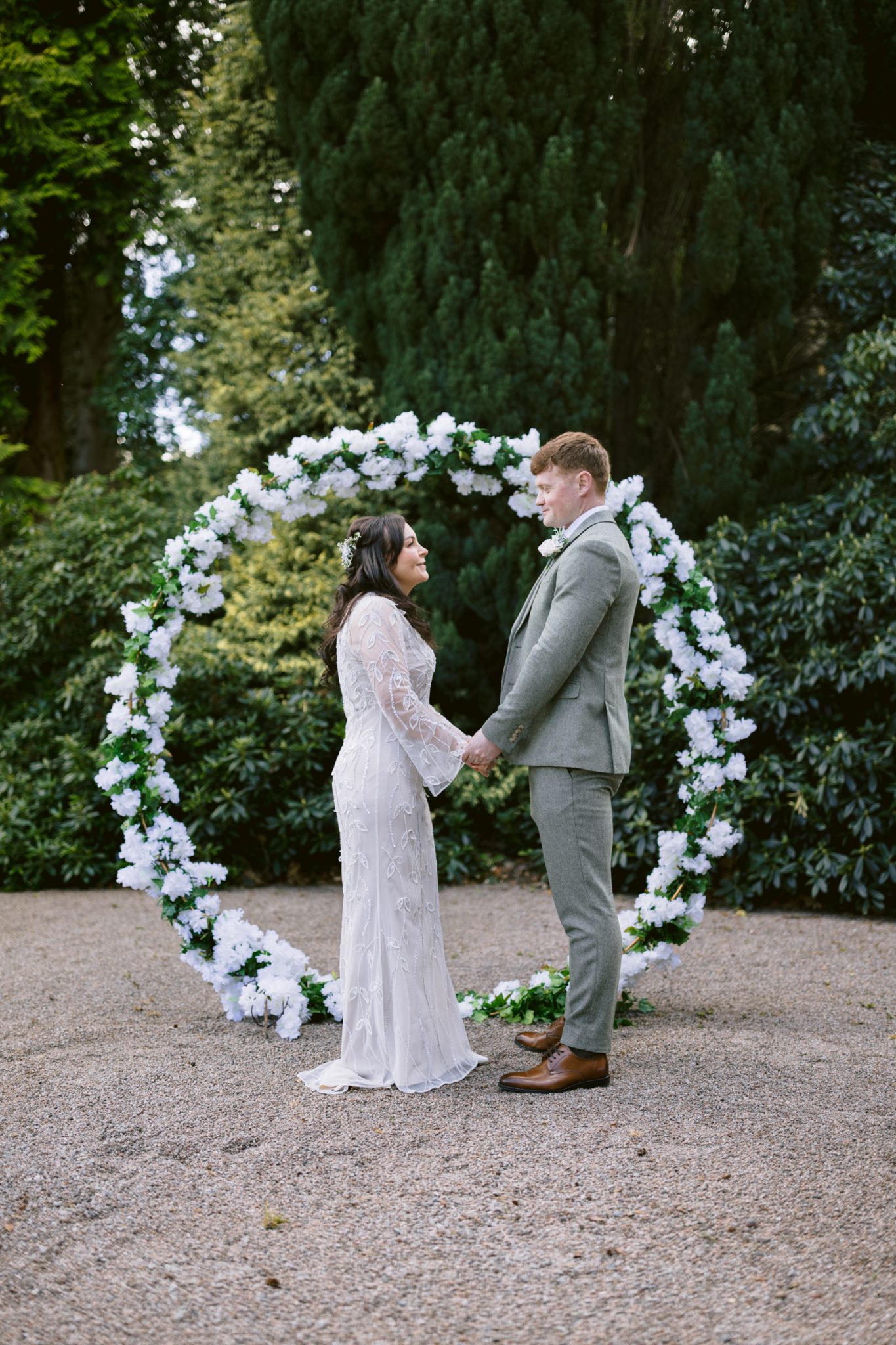 A couple in wedding attire stands holding hands in front of a circular floral arch outdoors, surrounded by greenery.