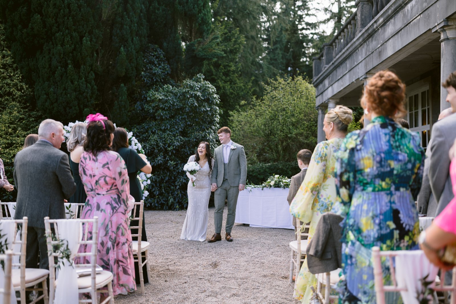 A bride and groom walk down the aisle together outdoors while wedding guests, some standing and some seated, watch on either side.