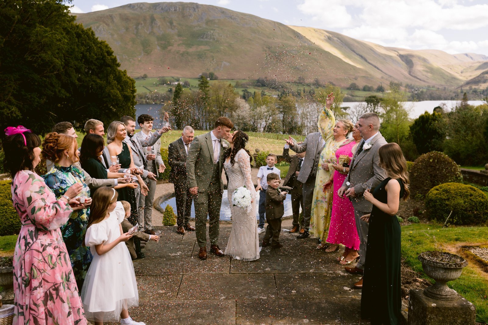 A bride and groom kiss outdoors as guests in formal attire throw confetti. Hills, trees, and a lake are visible in the scenic background.