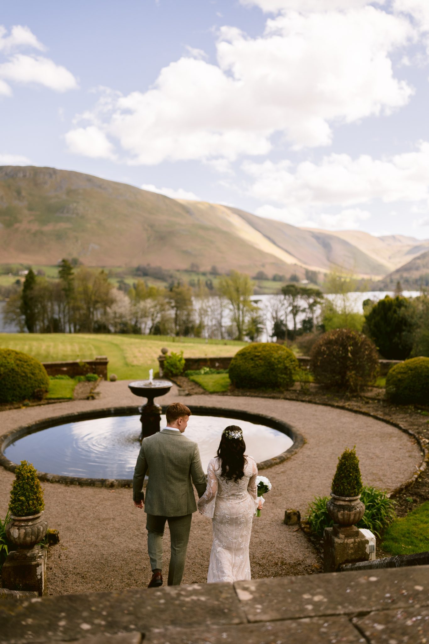 A bride and groom walk hand in hand toward a round pond in a landscaped garden, with a lake and mountains in the background under a partly cloudy sky.
