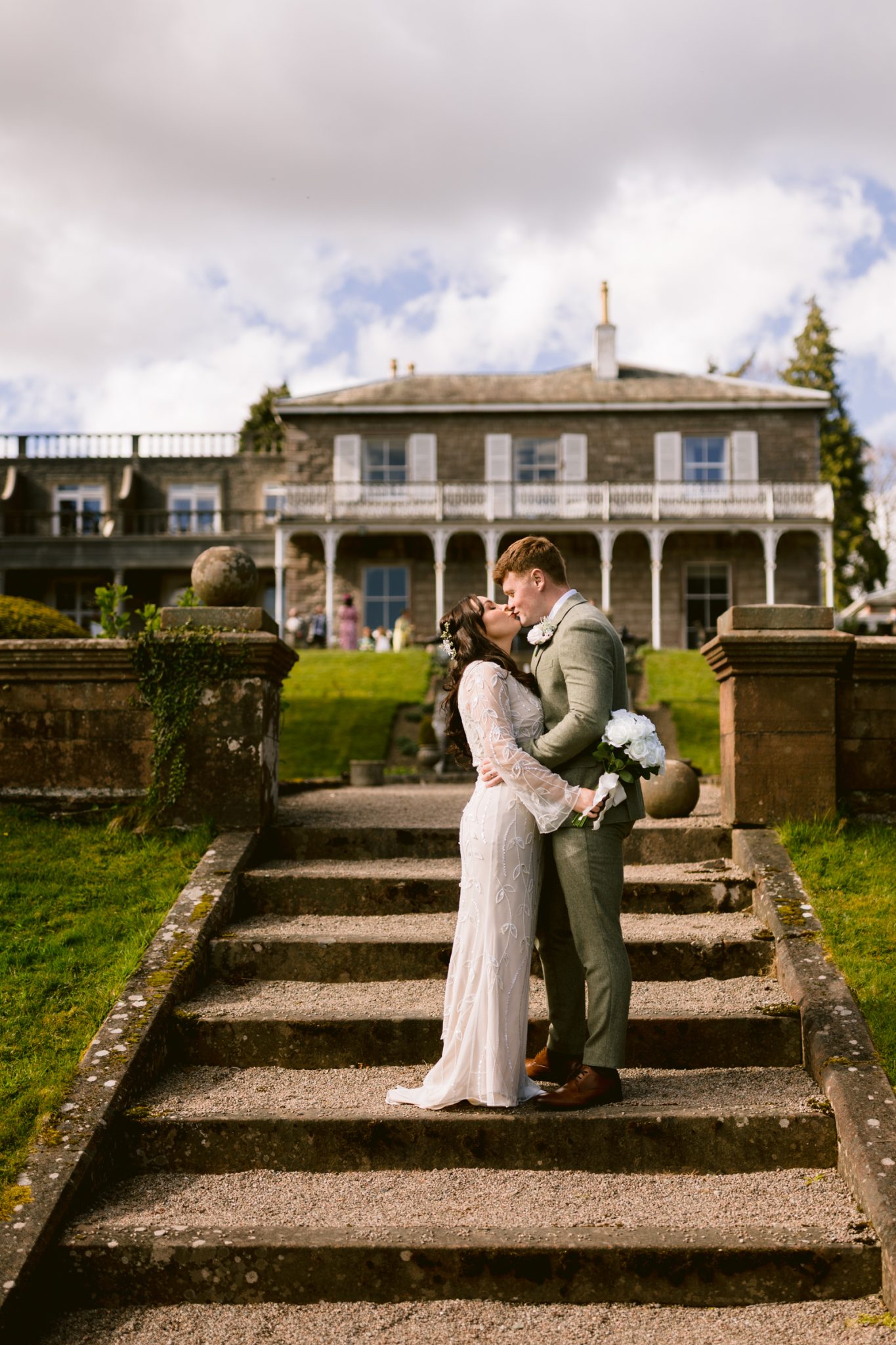 A bride and groom kiss on outdoor stone steps, holding a bouquet, with a large white house and manicured lawn in the background.