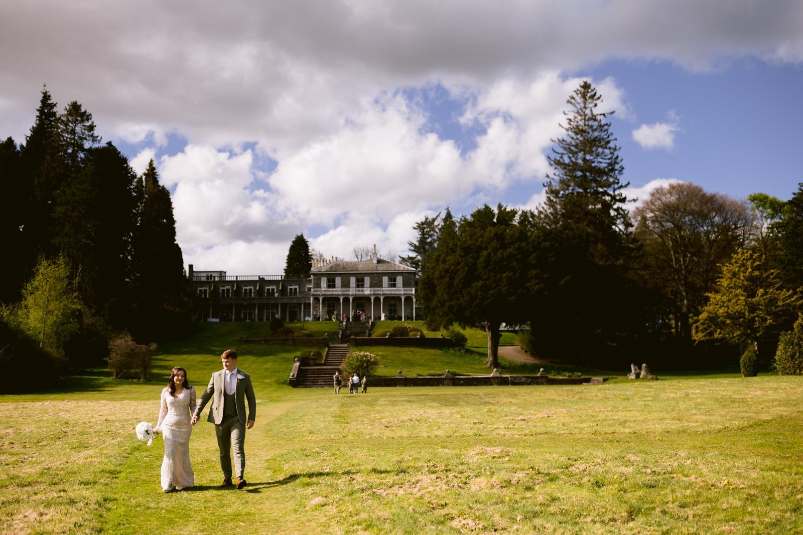 A bride and groom walk hand-in-hand across a grassy field in front of a large, historic building surrounded by trees under a partly cloudy sky.