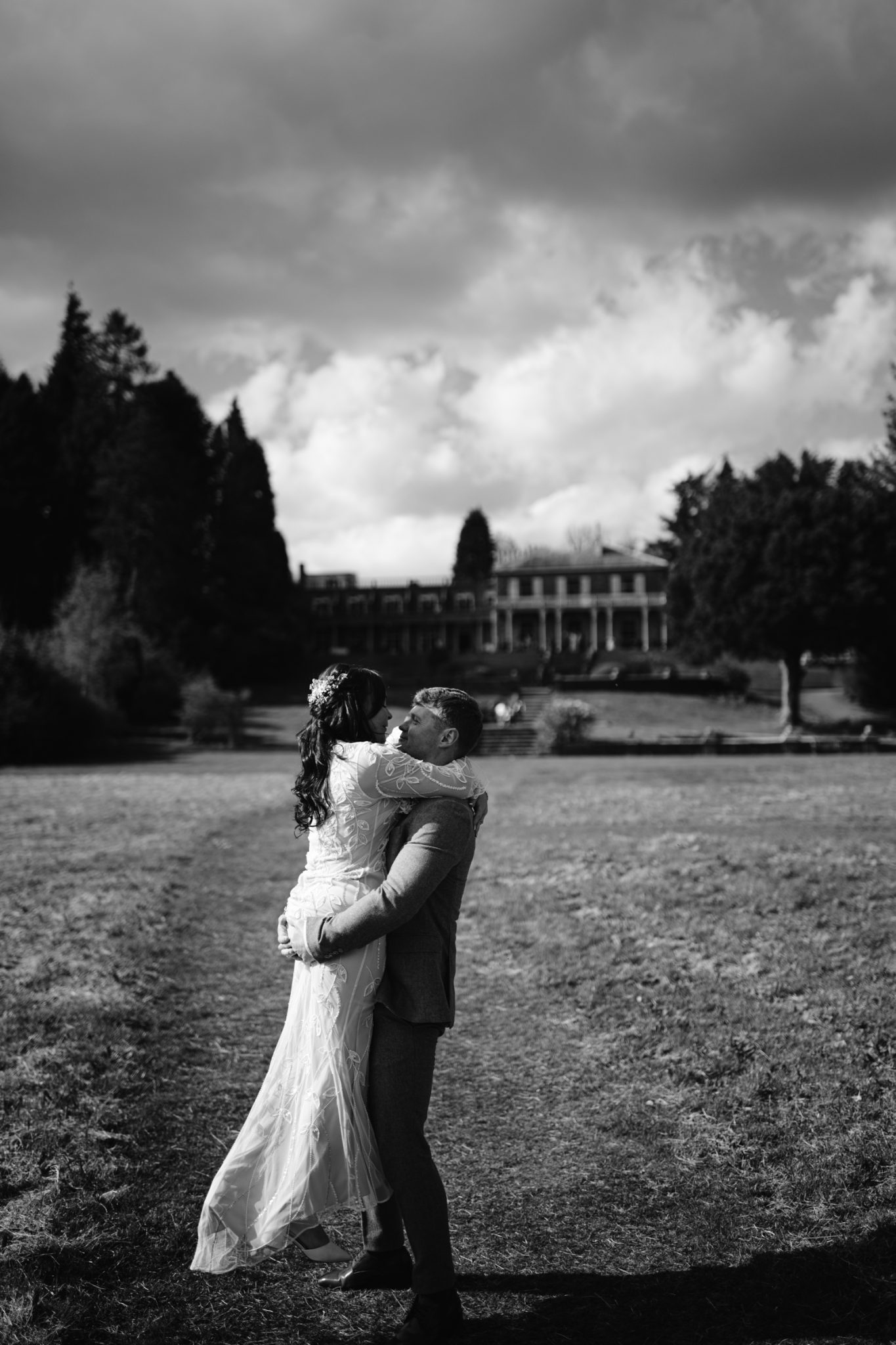 A man lifts a woman in a wedding dress outdoors on a grassy path, with a large building and trees in the background under a cloudy sky.