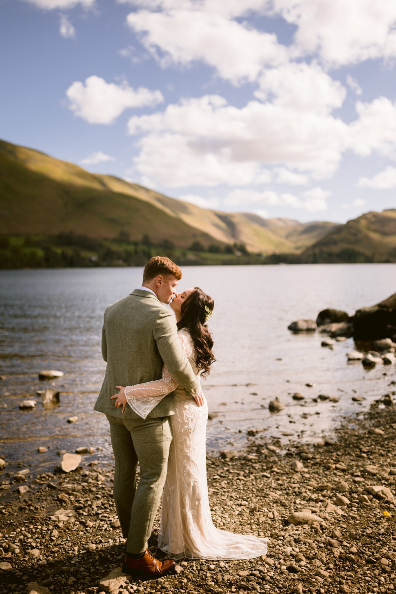 A bride and groom stand on a rocky lakeshore, embracing, with mountains and a partly cloudy sky in the background.
