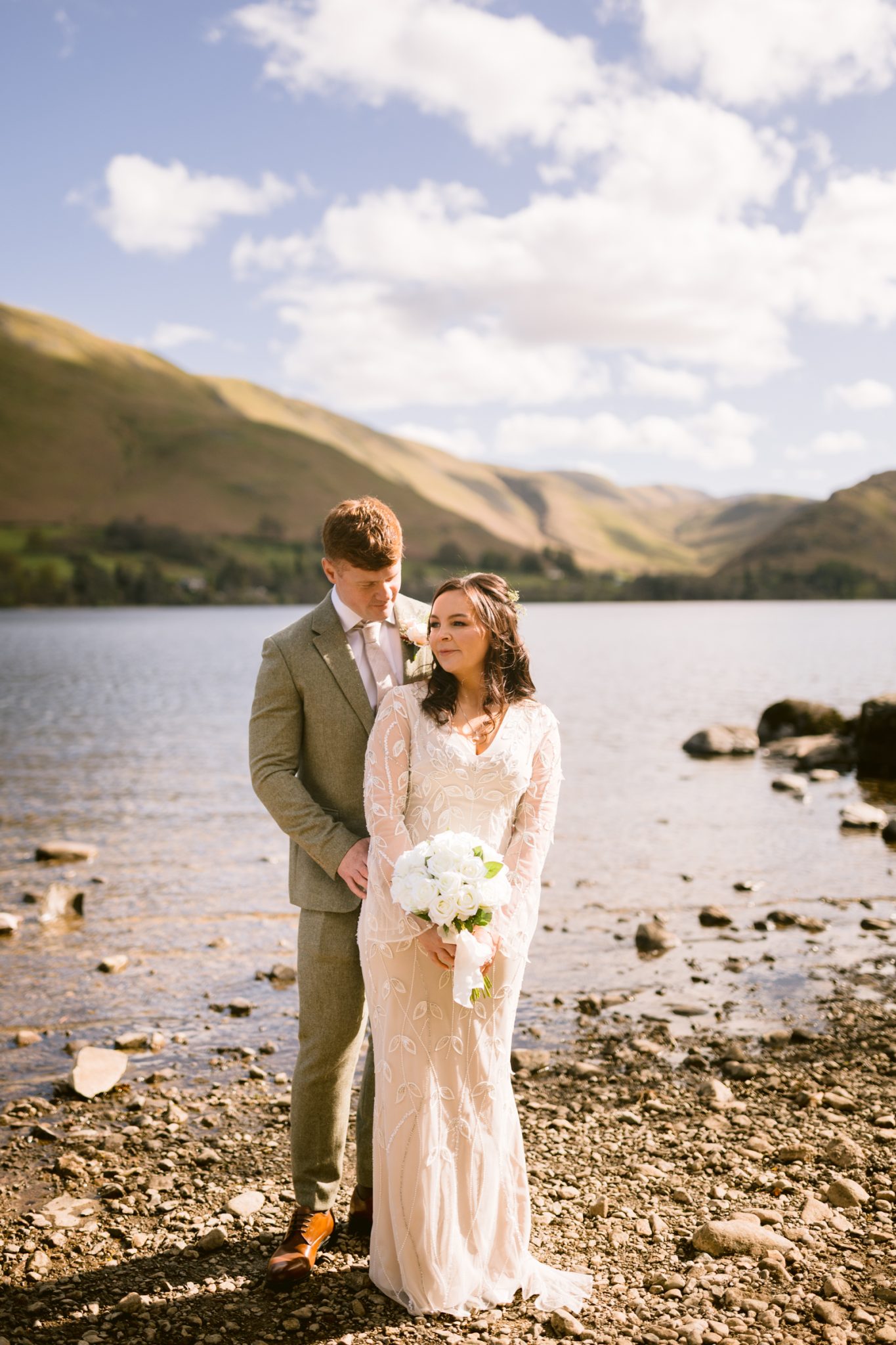 A bride and groom stand on a rocky lakeshore with mountains and a partly cloudy sky in the background. The bride holds a bouquet of white flowers.