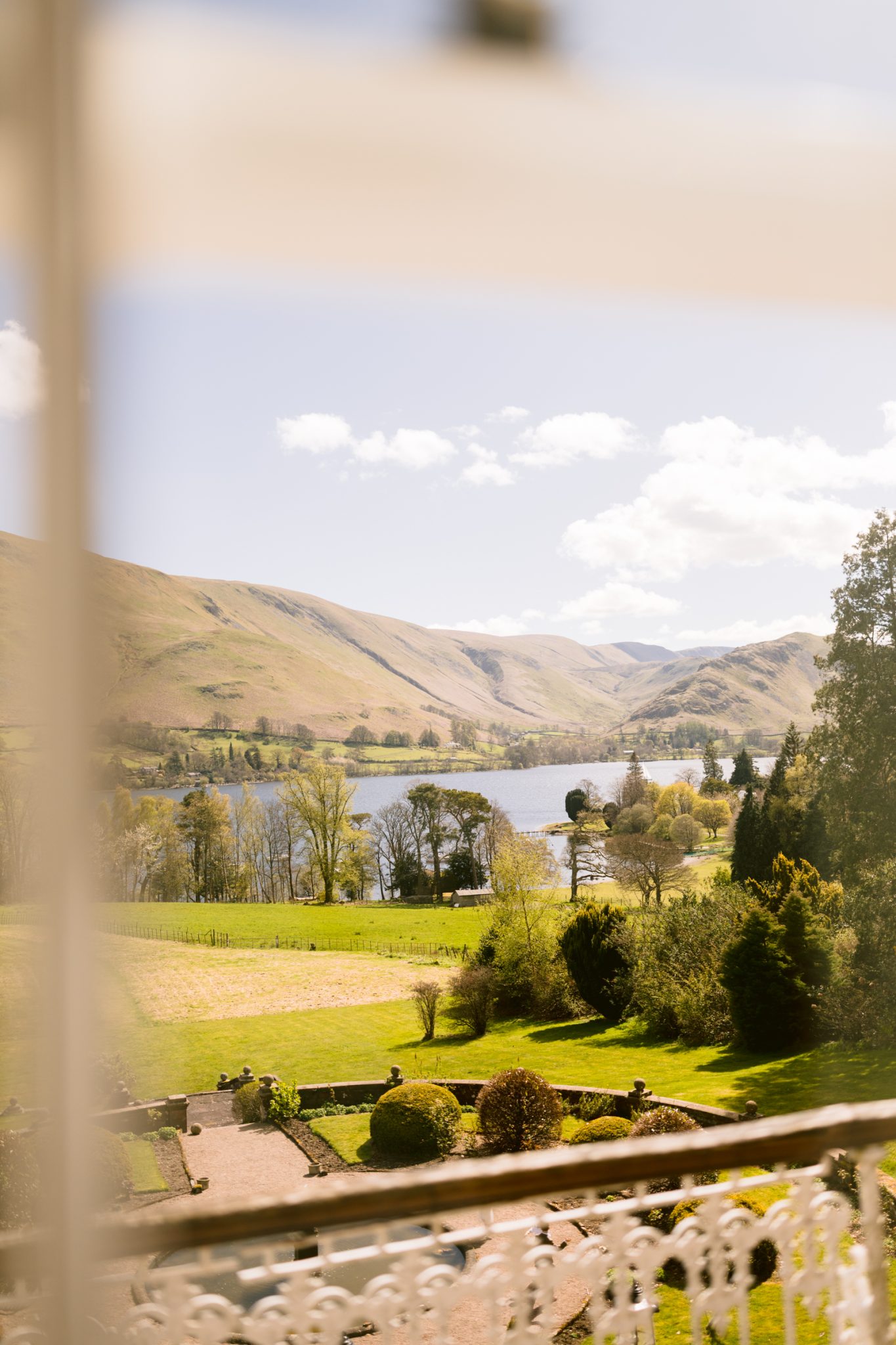 View of a green valley with scattered trees, a river or lake, distant hills, and a clear sky, seen from a balcony with part of the railing visible in the foreground.