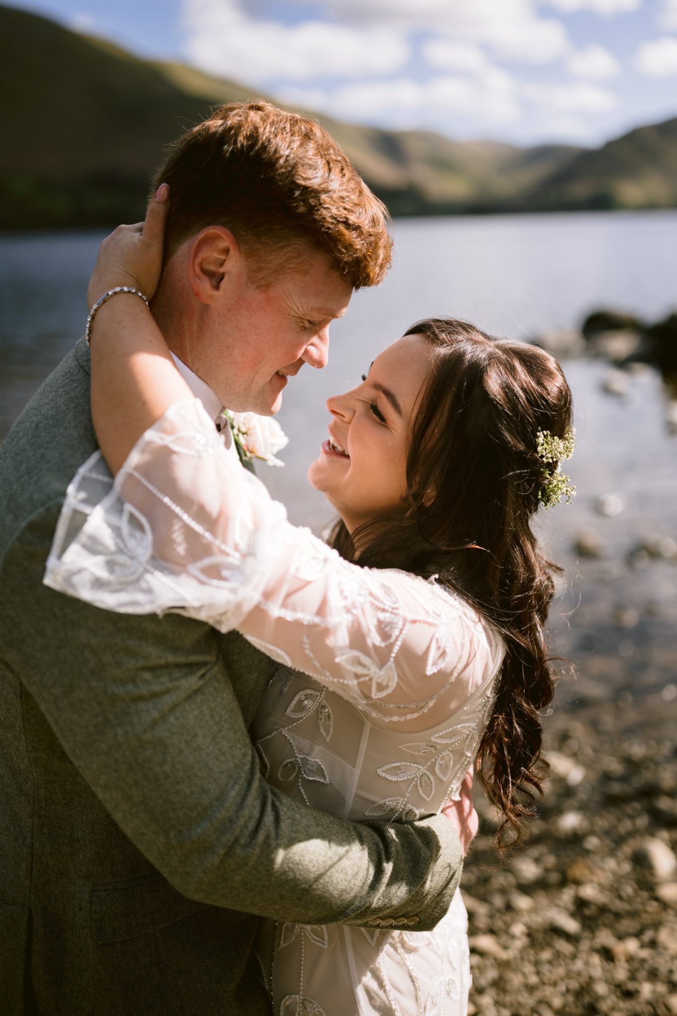 A couple in wedding attire embraces and smiles at each other by a lakeside with hills in the background on a sunny day.