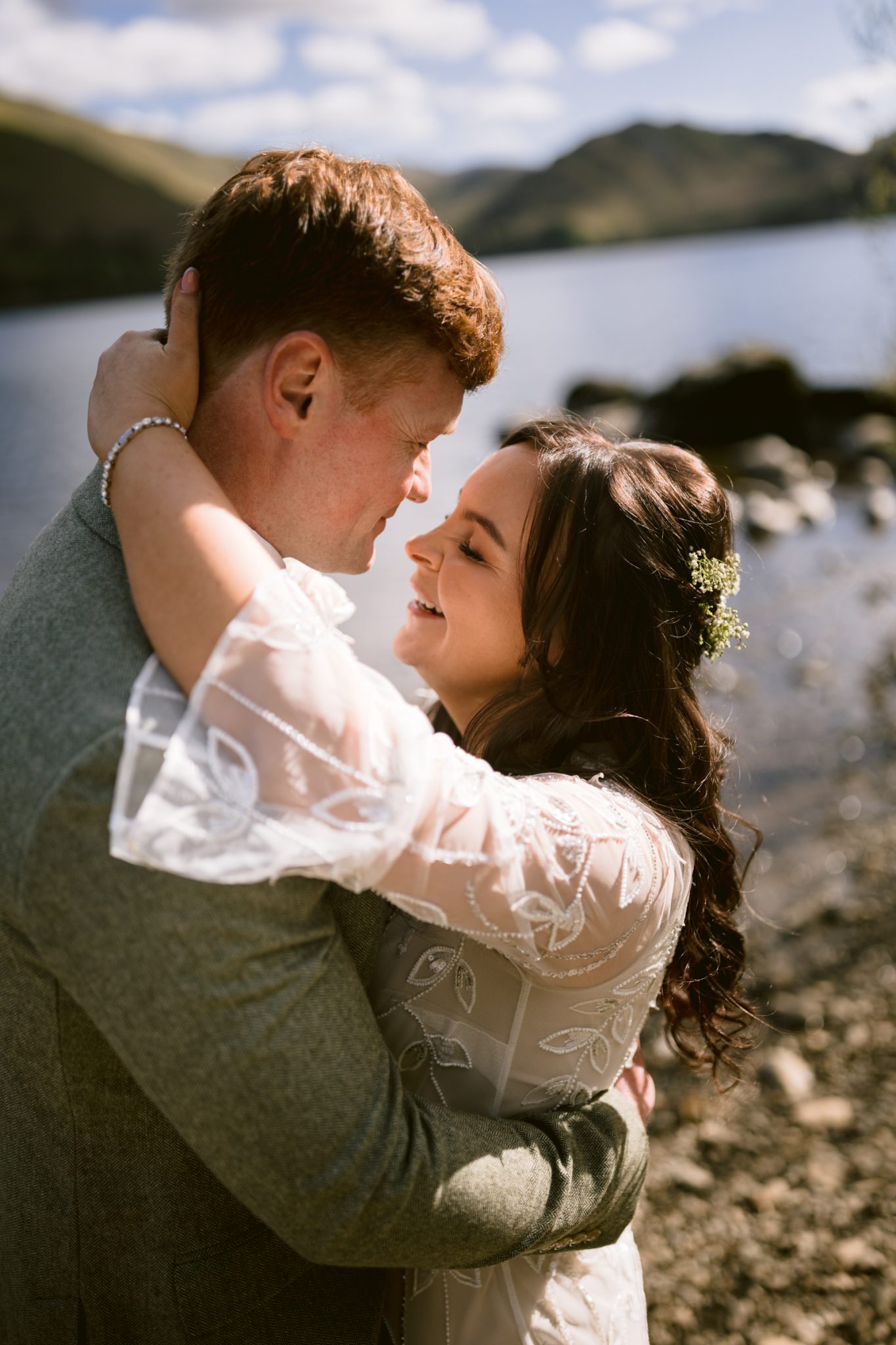 A couple embraces and smiles at each other by a lakeside, with mountains and blue sky in the background.