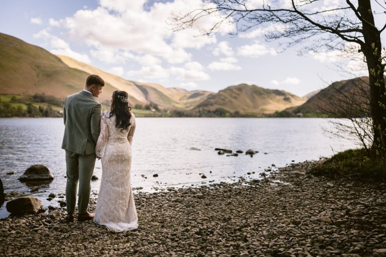 A couple in wedding attire stands on a rocky lakeshore, facing the water and distant hills under a partly cloudy sky.
