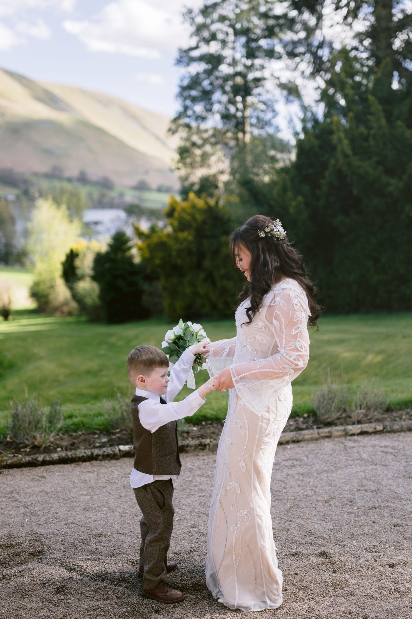 A woman in a white dress holds hands with a young boy in a vest and bow tie on a gravel path with green trees and hills in the background.