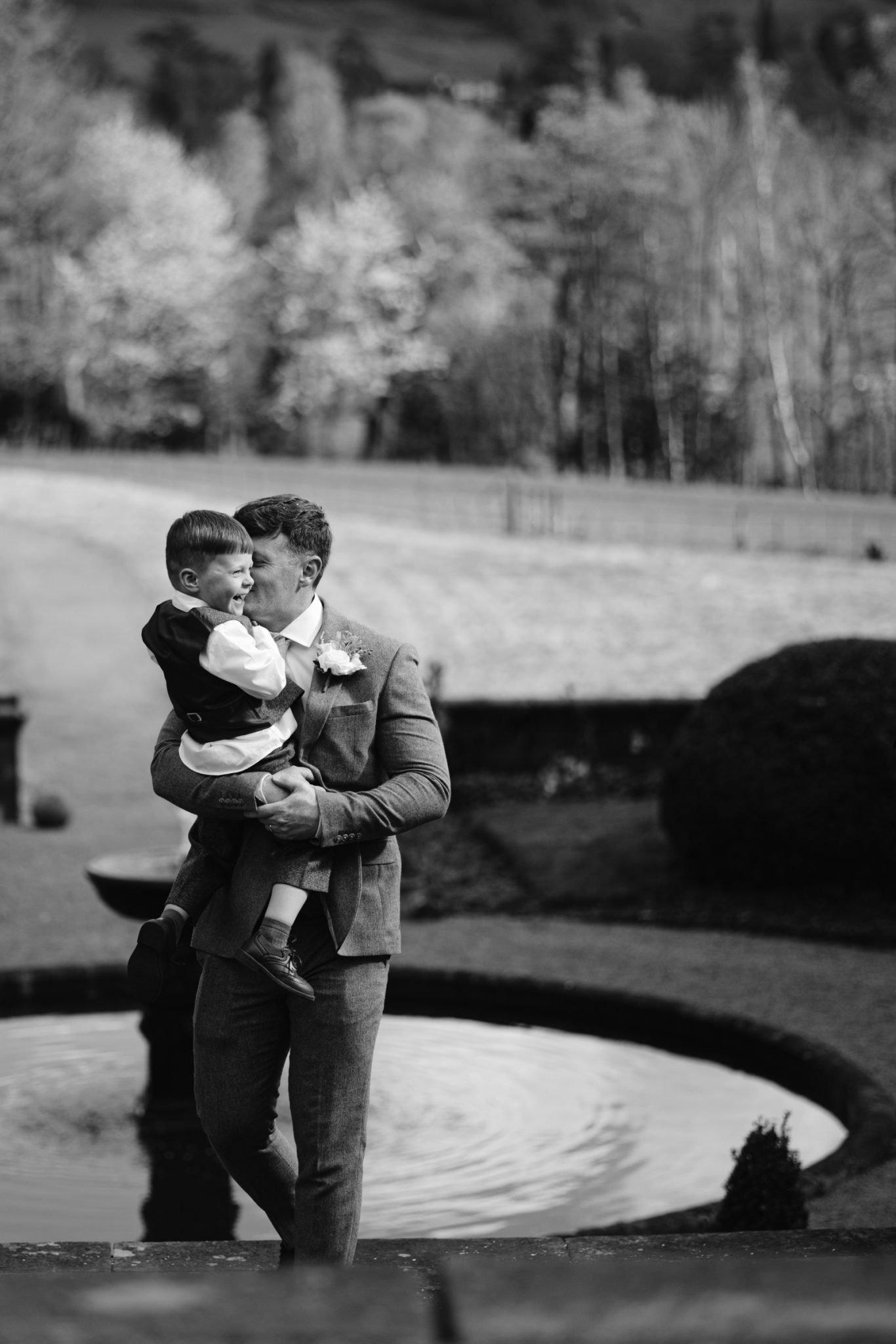A man in a suit holds a young boy in his arms while standing near a circular pond in an outdoor garden setting.