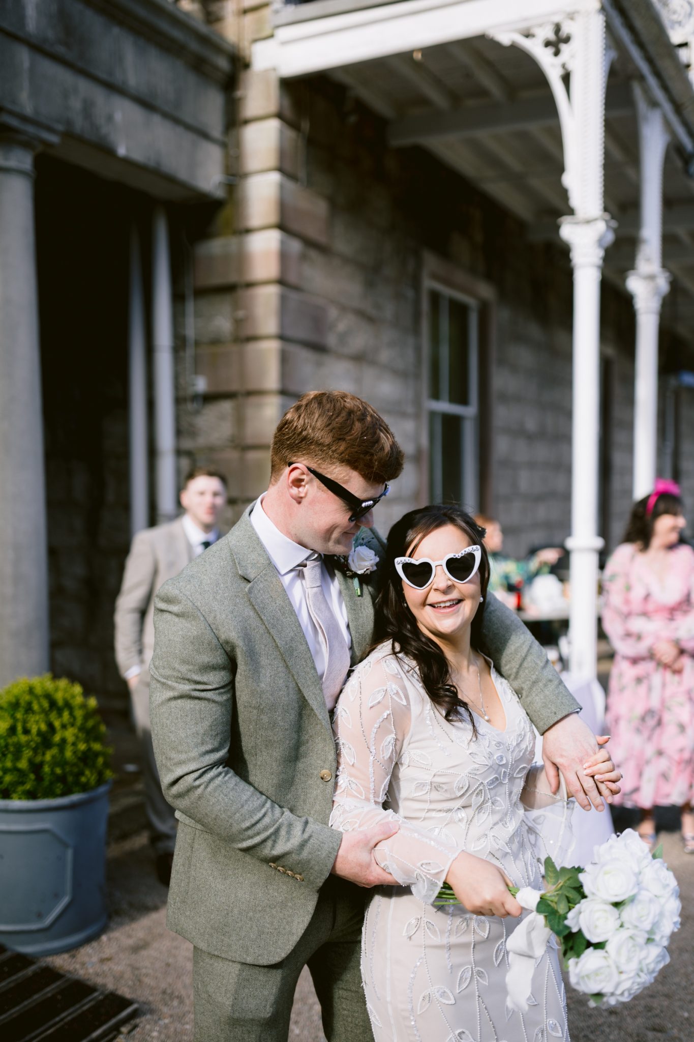 A couple dressed in wedding attire, both wearing sunglasses, pose outdoors in front of a stone building with white columns; the bride holds a bouquet of white flowers.