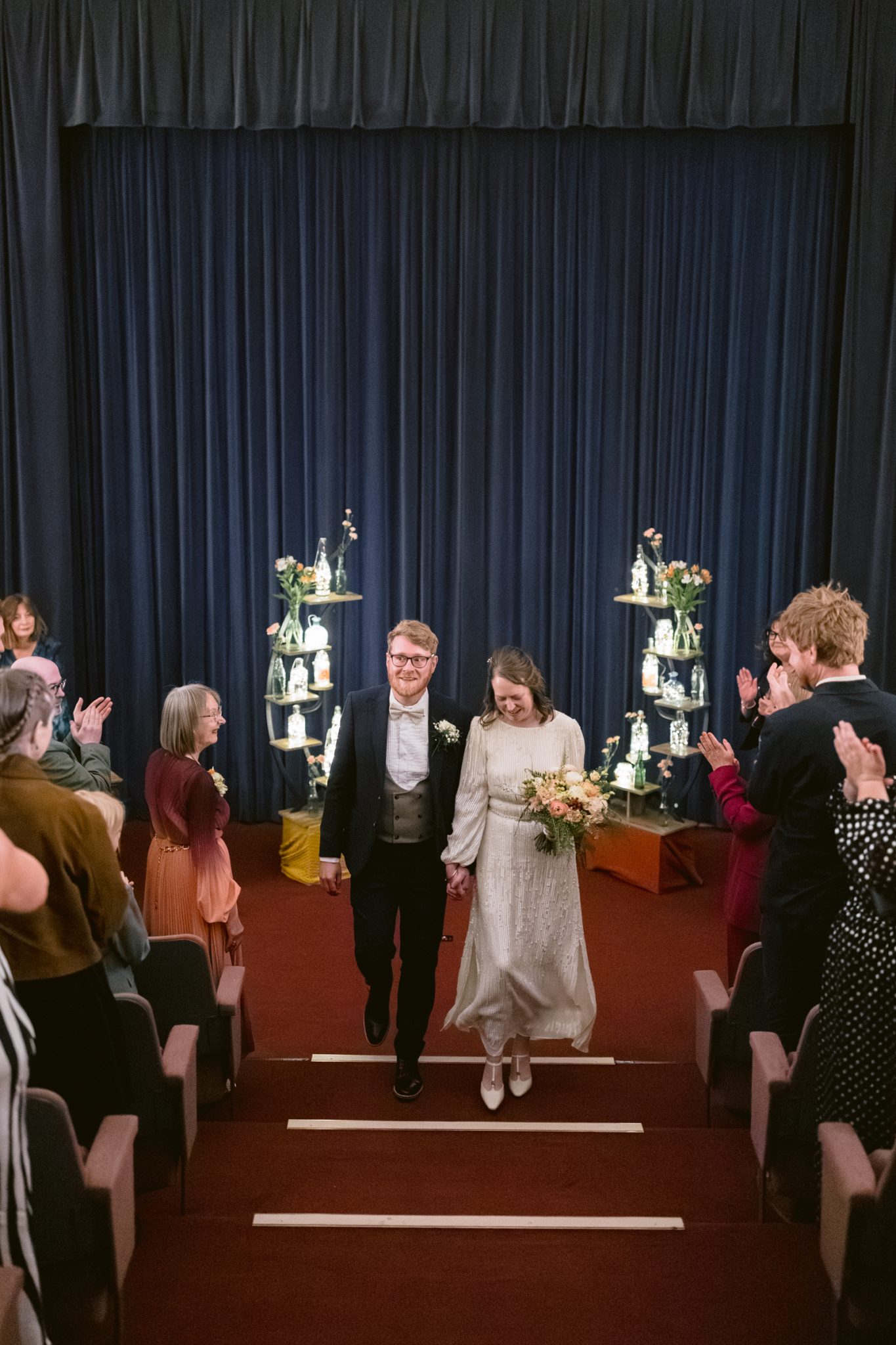 A bride and groom walk up the aisle together, holding hands and smiling, as guests applaud in a theater-like setting with floral arrangements on stage at their beautiful Tullie House wedding.
