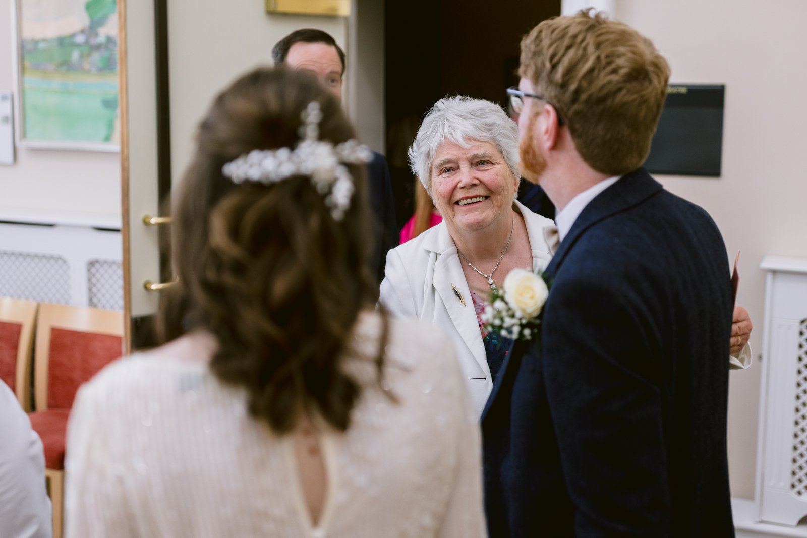 An older woman, smiling, stands indoors at a Tullie House Wedding, facing a man in a suit and a woman in a white dress with her back to the camera.