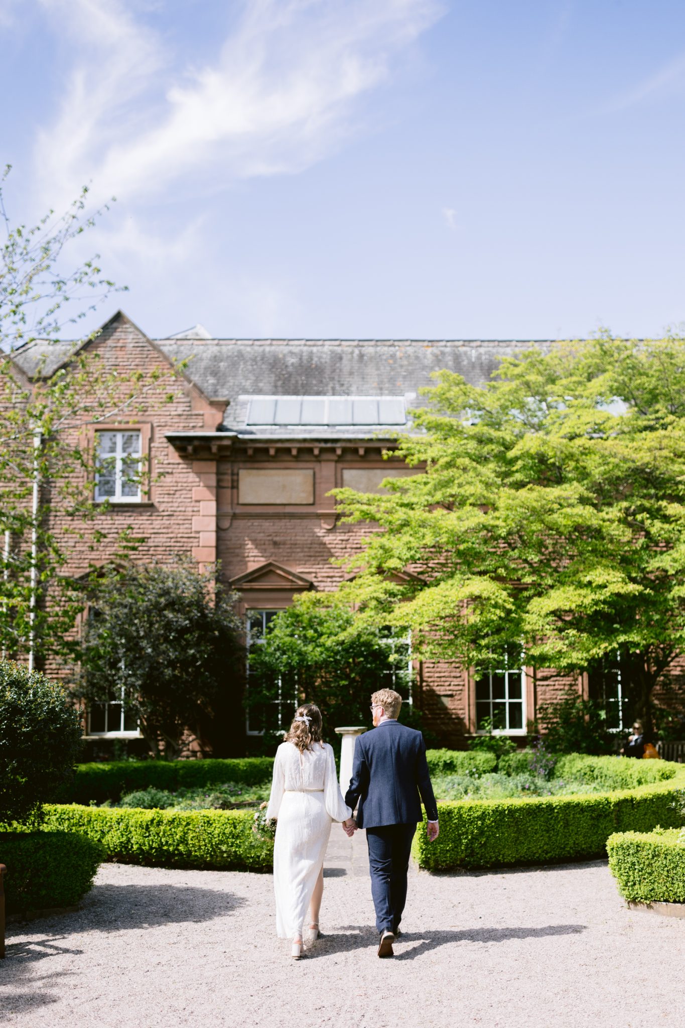 A couple dressed in formal attire walks hand in hand towards Tullie House, surrounded by greenery under a blue sky—a perfect scene for a romantic Spring wedding.