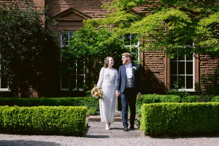 A bride and groom walk hand in hand through a garden at Tullie House, with lush greenery and a brick building setting the scene for their beautiful spring wedding.