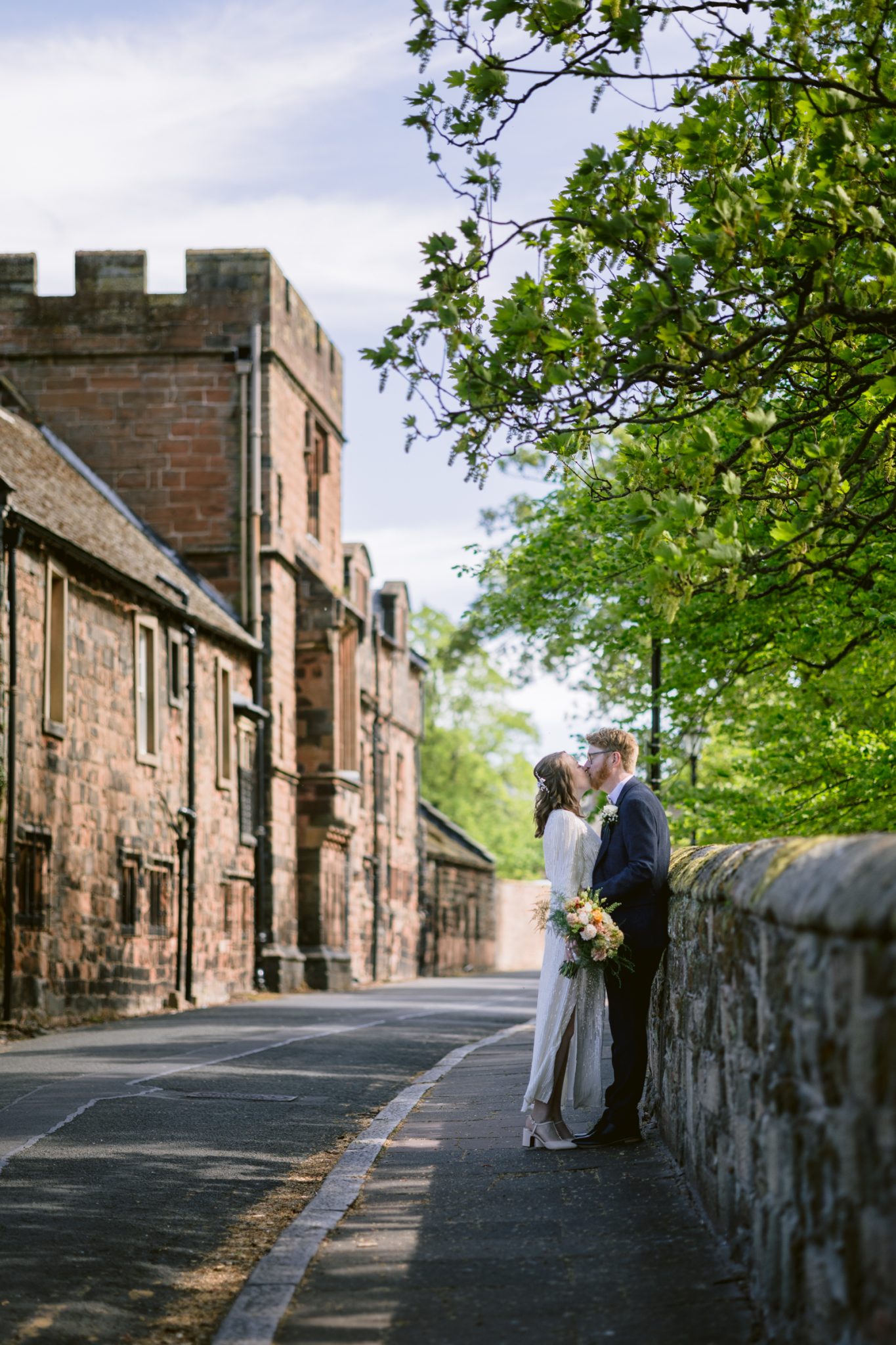 A couple in wedding attire stands kissing by a stone wall along a quiet street near Tullie House, surrounded by historic buildings and green trees—a perfect scene for a spring wedding.