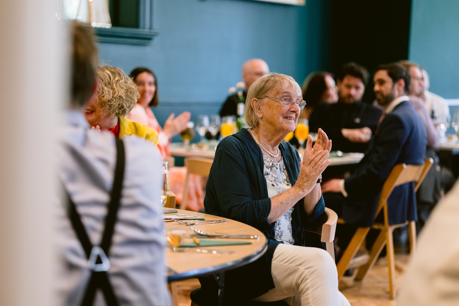 An older woman sits at a round table and claps her hands while other guests chat in the background at a joyful Tullie House Wedding gathering.