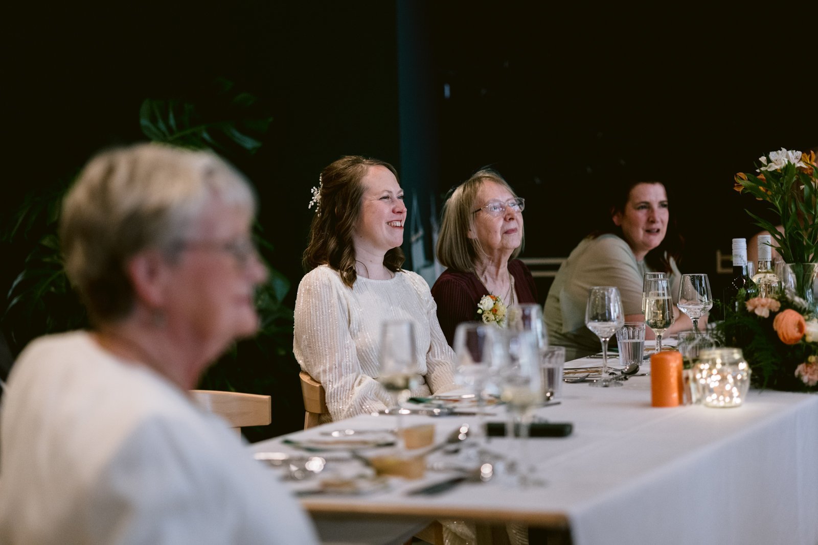 Four women sit at a decorated table, smiling and facing forward, with glasses and floral arrangements set in front of them, capturing the joy of a Spring wedding at Tullie House.