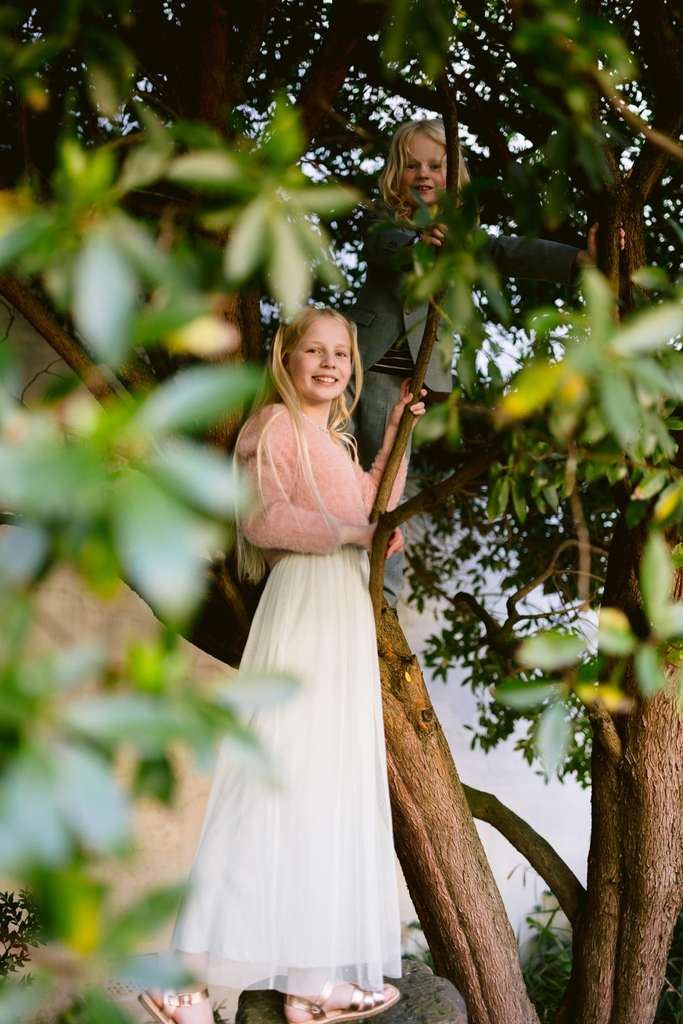 Two children, a girl in a long white skirt and pink top and a boy in a gray shirt, are climbing and standing in a tree surrounded by green leaves at Tullie House, capturing the joyful spirit of a spring wedding.