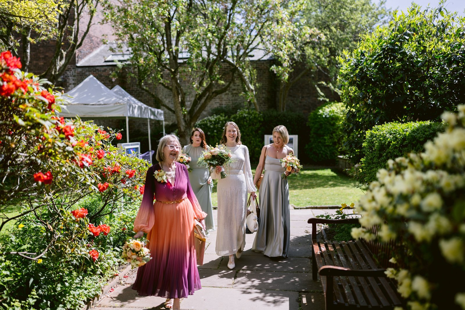 Four women dressed in formal attire, holding bouquets, walk along a garden path on a sunny day surrounded by greenery and flowers—capturing the joyful spirit of a Spring Wedding.