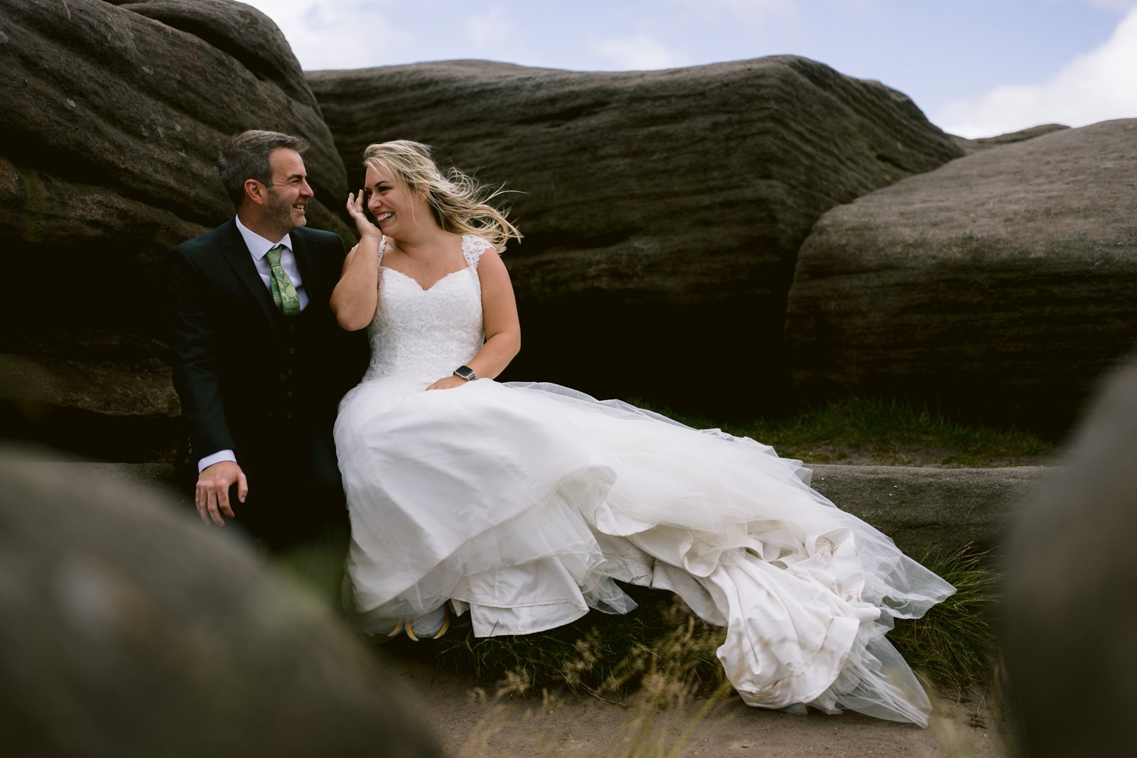 A bride in a white gown and a groom in a black suit sit together on rocks outdoors, smiling and laughing.