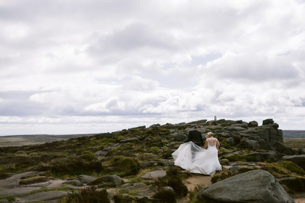 A bride and groom walk together on a rocky, grassy hill under a cloudy sky, with the bride’s dress trailing behind.