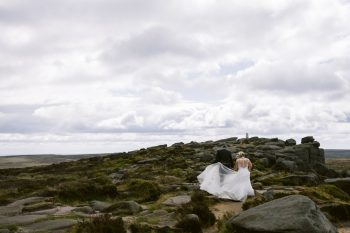 A bride and groom walk together on a rocky, grassy hill under a cloudy sky, with the bride’s dress trailing behind.