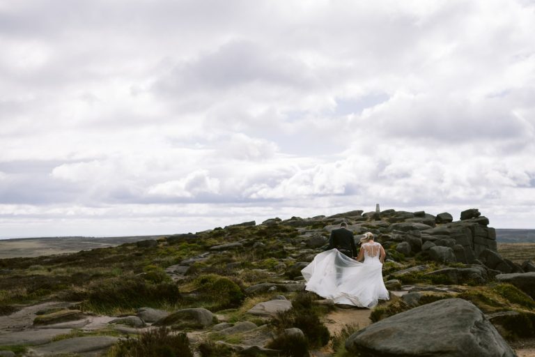 A bride and groom walk together on a rocky, grassy hill under a cloudy sky, with the bride’s dress trailing behind.
