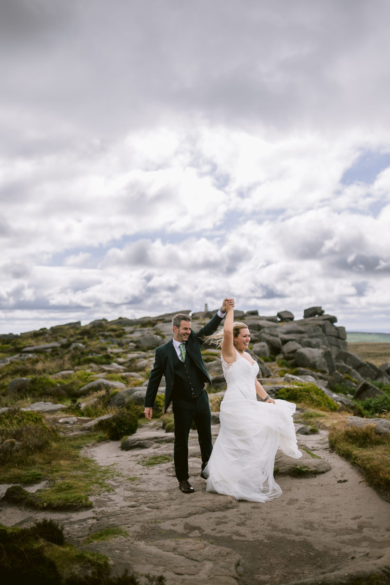 A bride and groom walk on a rocky path outdoors, with the groom holding up the bride’s hand. The bride wears a white dress and the groom wears a suit. The sky is cloudy.