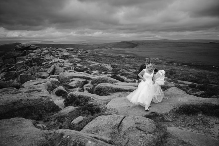 A couple in wedding attire walk across a rocky landscape under a cloudy sky, with hills and open fields in the background.