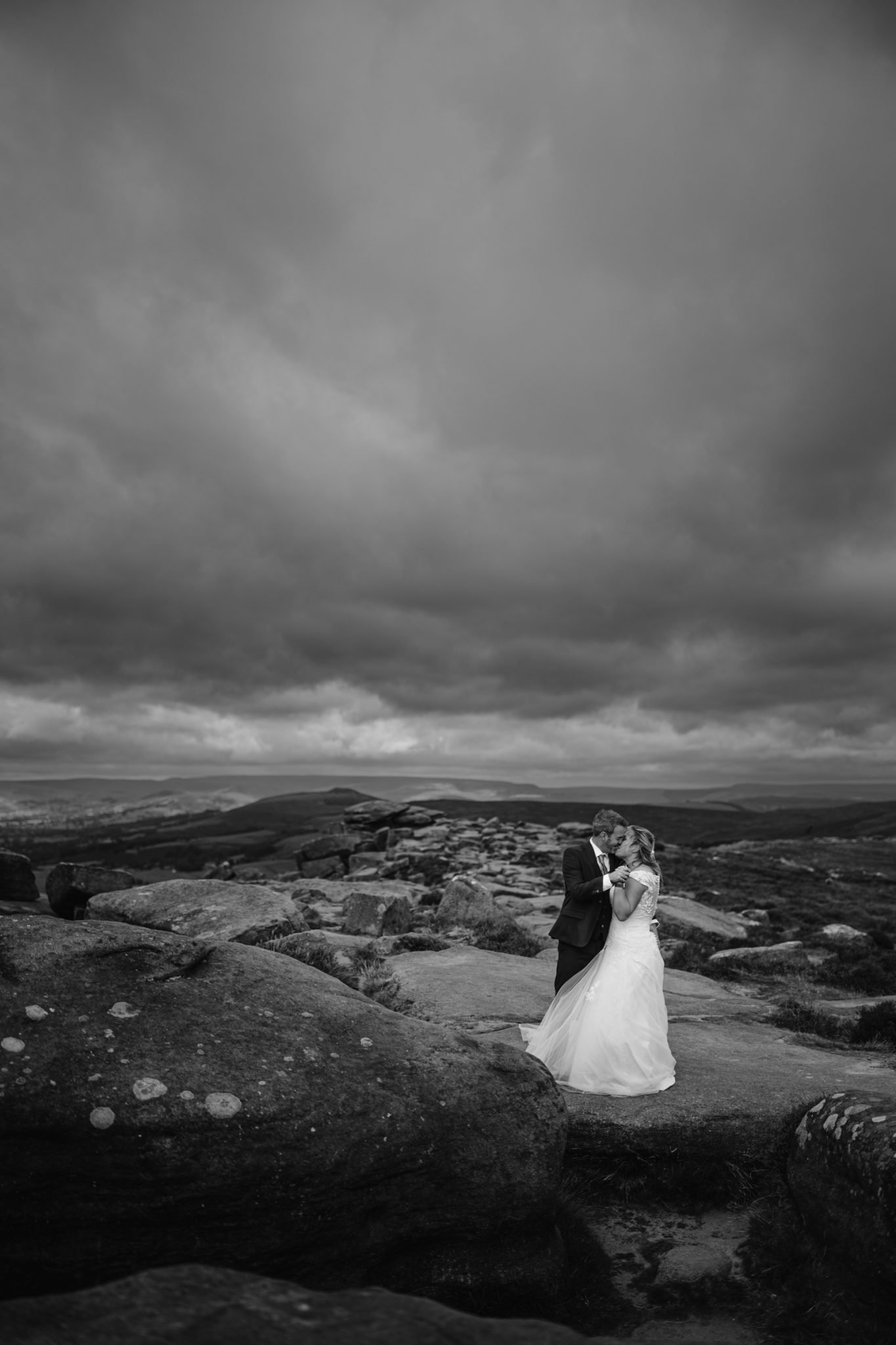 A couple in wedding attire embraces on rocky terrain under a cloudy sky, with distant hills in the background.