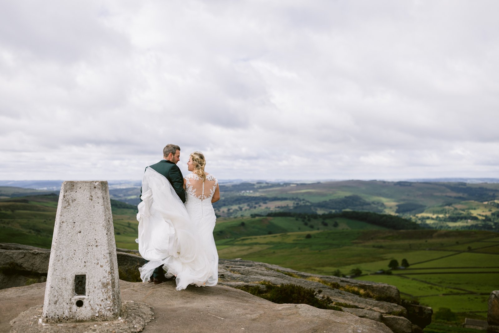 A bride and groom stand on a rocky hilltop overlooking a vast, green landscape under a cloudy sky, with a concrete survey marker in the foreground.