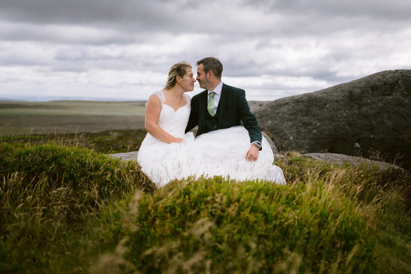 A bride and groom sit close together outdoors on grass and rocks, dressed in wedding attire, under a cloudy sky.